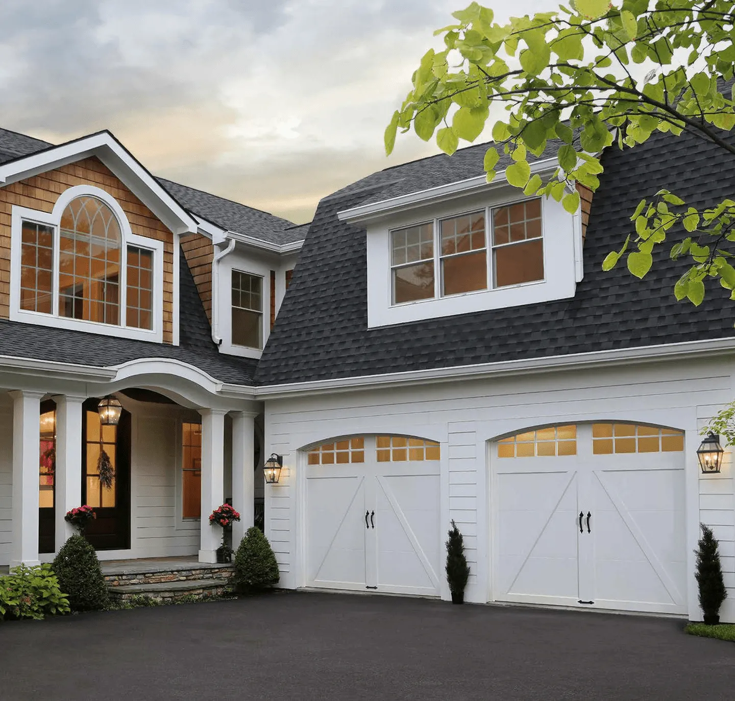 White two-car carriage-house garage doors with arched windows on a shingle-style home at dusk